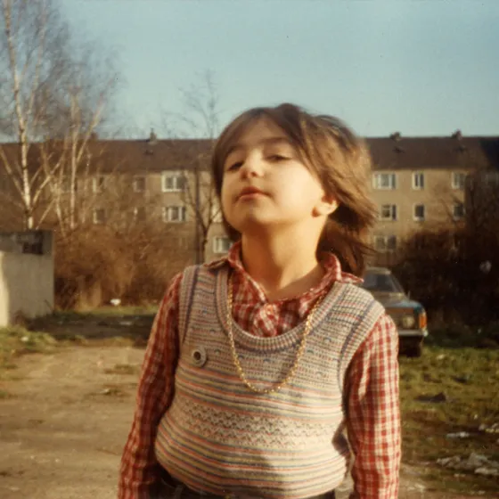 Die siebenjährige Çiğdem Satır, mit kariertem Hemd, Pullunder und Kette steht auf einem Hof in Duisburg und schaut mit einem verwegenen Lächeln in die Kamera. Im Hintergrund sind Wohnhäuser, einige Birken und eine Plakatwand zu erkennen.