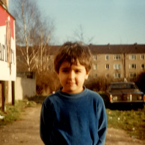 Der fünfjährige Ümit Satır steht in einem blauen Pullover Satır auf einem Hof in Duisburg und schaut mit einem etwas schüchternen Lächeln in die Kamera. Im Hintergrund sind Wohnhäuser, einige Birken und eine Plakatwand zu erkennen.