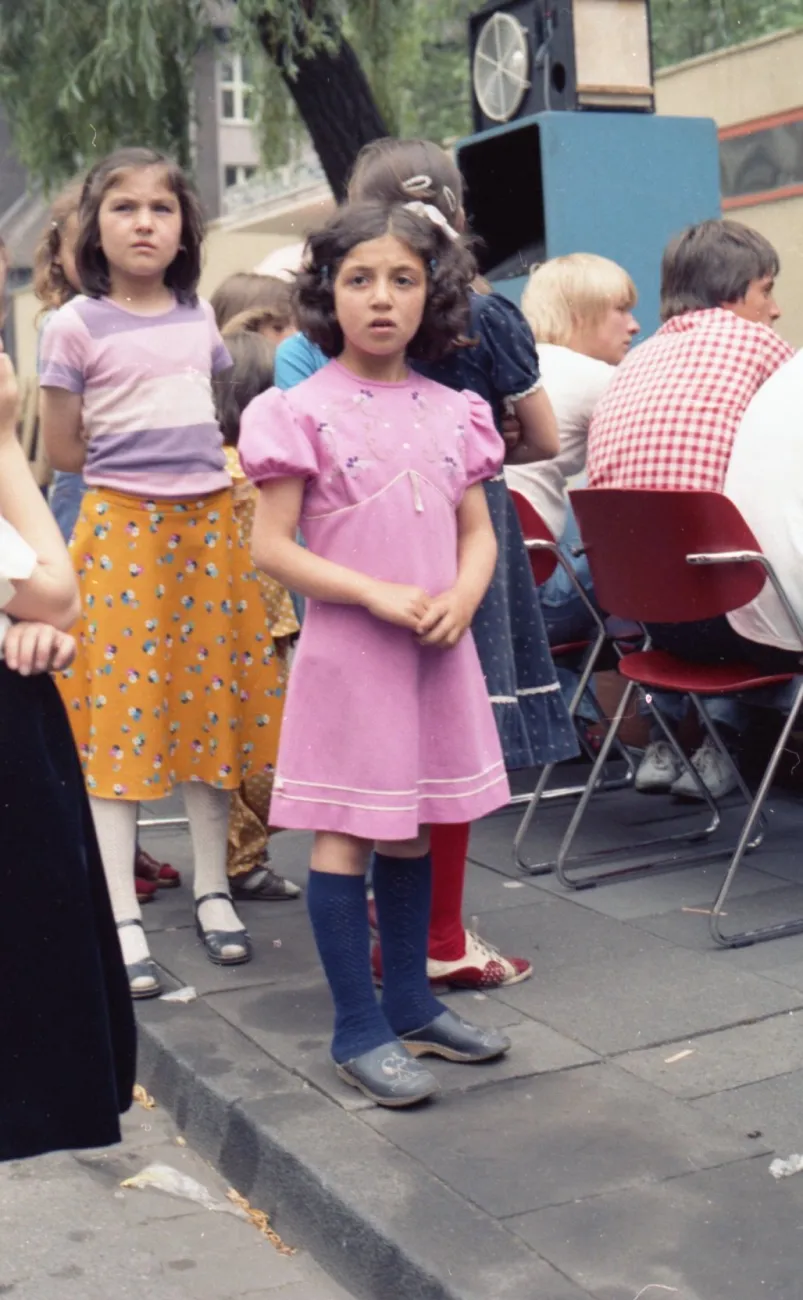 In der Mitte des Bildes stehen Mädchen im Alter von 5-8 Jahren. Im Vordergrund ist ein Mädchen in einem rosa Kleid, die direkt in die Kamera schaut. Dahinter sind Stuhlreihen mit Personen zu erkennen, die der Kamera den Rücken zugedreht haben.