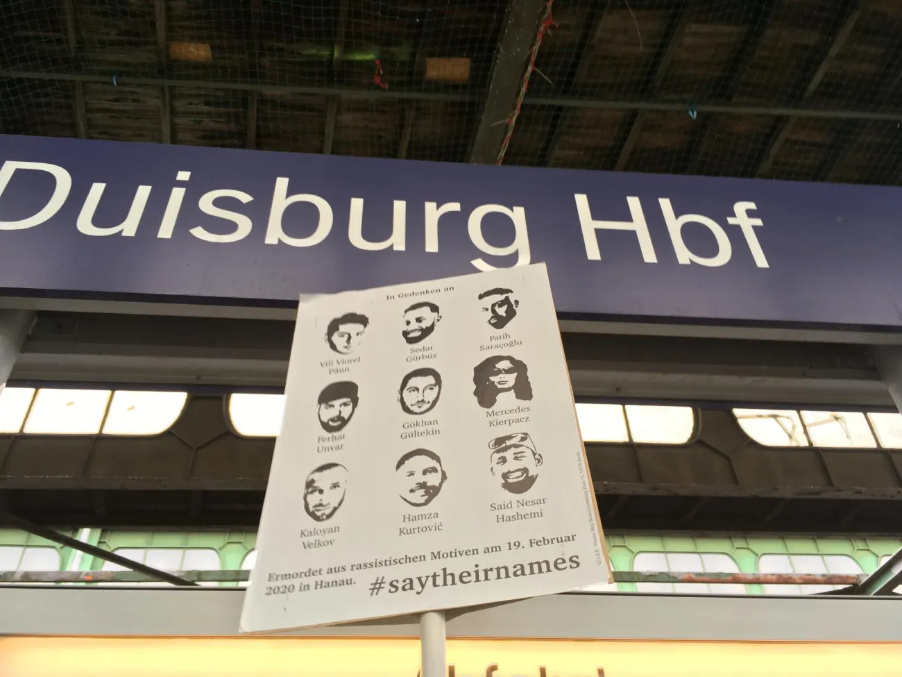 Ein Plakat im Gedenken an die Opfer des rassistischen Anschlags vom 19.02.2020 in Hanau vor einem Bahnhofsschild mit der Aufschrift Duisburg Hbf.