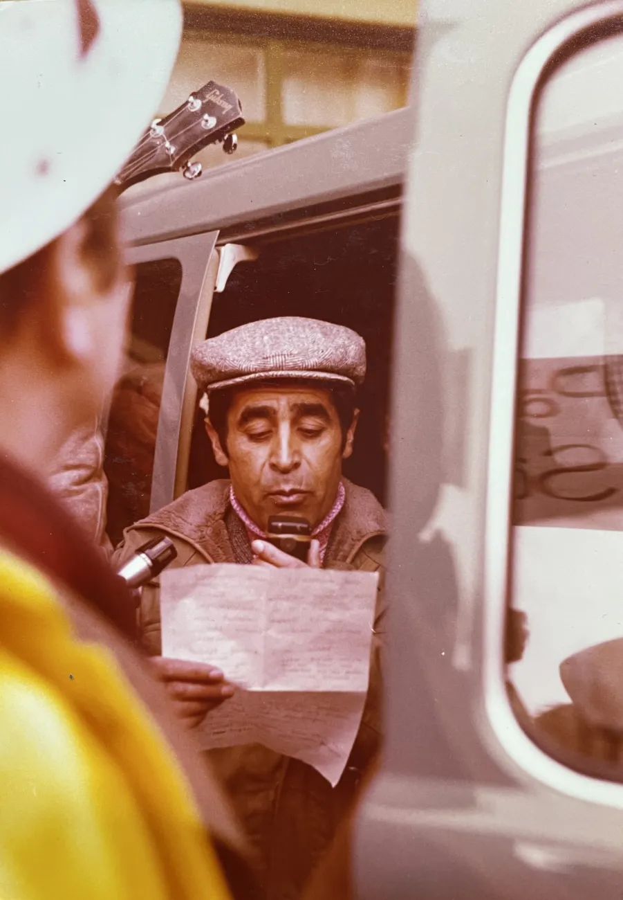 Historische Farbfotografie einer öffentlichen Ansprache: Ein Mann steht in der Tür eines Fahrzeugs und verliest mit einem Handmikrofon eine Erklärung von einem Blatt Papier.